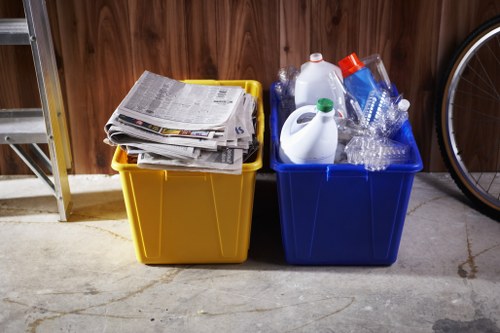 Two plastic recycling bins placed side by side on a concrete floor in a garage or storage area, with a wooden wall in the background. The bin on the left is yellow and filled with newspapers and folded paper waste, while the bin on the right is blue and contains clear plastic bottles, some with caps, and a white plastic bottle with a handle. The bins are positioned near a metal shelving unit and a bicycle wheel is partially visible on the far right. The scene reflects a typical waste collection setup for household recycling, aligned with rubbish removal services in Bromley. The lighting is natural and the environment appears tidy, emphasizing the organization of recyclable materials awaiting collection or disposal.